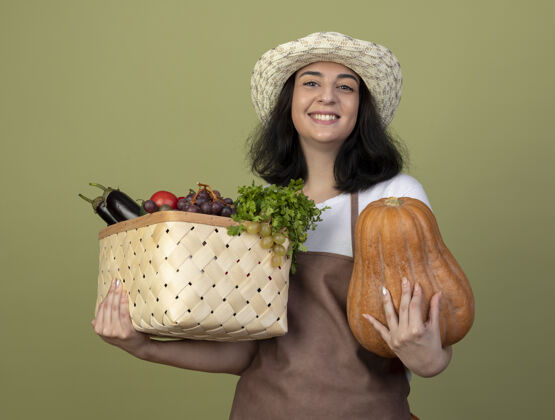 人微笑着的年轻黑发女园丁穿着制服 戴着园艺帽 把菜篮子和南瓜隔离在橄榄绿的墙上黑发橄榄微笑