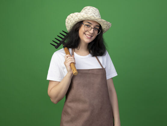 帽子微笑着的年轻黑发女园丁戴着眼镜和制服 戴着园艺帽 肩上扛着耙子 隔离在绿色的墙上光学人姿势