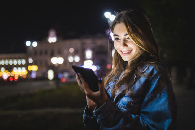女人年轻快乐的女人用平板电脑在户外模糊的夜晚城市灯光聊天时尚微笑