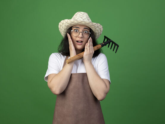 年轻人焦急的年轻黑发女园丁戴着眼镜和制服戴着园艺帽把手放在脸上拿着耙子隔离在绿色的墙上帽子手持女性