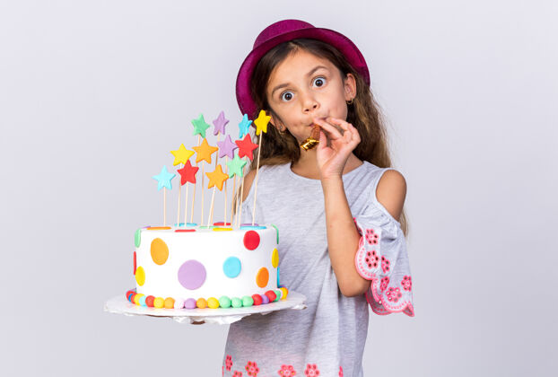孤立惊讶的小白种女孩 戴着紫色派对帽 拿着生日蛋糕 吹着派对口哨 隔离在白色墙壁上 留着复制空间派对吹小