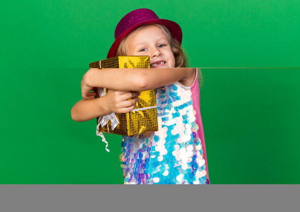 帽子微笑的金发小女孩 戴着紫色派对帽 拥抱着礼品盒 隔离在绿色墙壁上 有复制空间孤立女孩拥抱