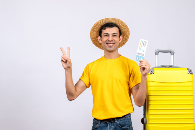 持有正面图：年轻游客站在黄色手提箱旁 拿着机票做v形标志年轻游客人帽子
