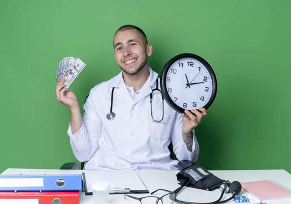 脸面带微笑的年轻男医生穿着医用长袍和听诊器坐在办公桌旁 工作工具把时钟和钱隔离在绿色的墙上姿势时钟抱着