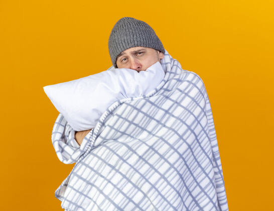 斯拉夫苍白的年轻金发病男子戴着冬天用格子布包裹的帽子抱住隔离在橙色墙上的枕头格子人表情