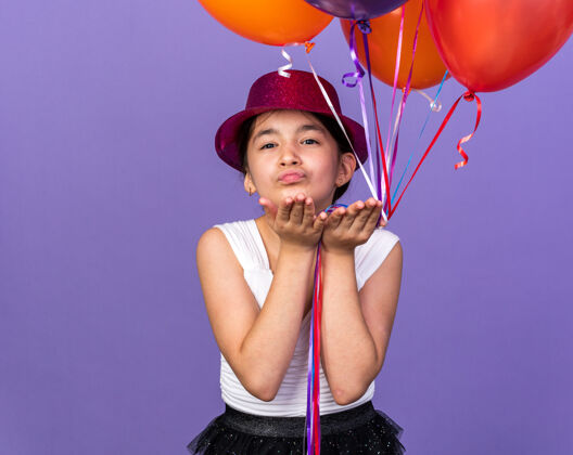 手戴着紫色派对帽 手持氦气球 手孤立在紫色墙壁上 留有复制空间的年轻白种人女孩高兴地送上了吻发送女孩孤立
