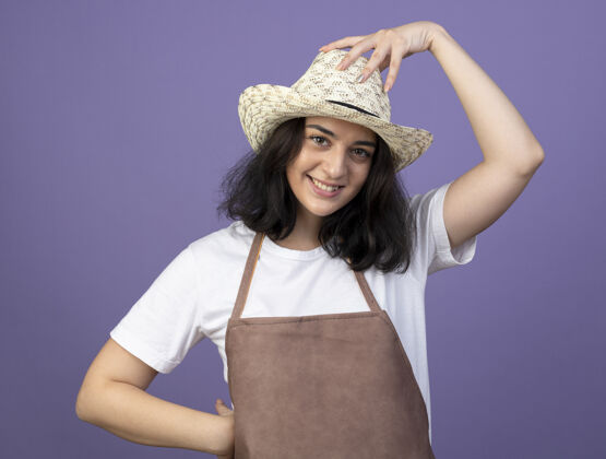 手微笑着的年轻黑发女园丁穿着制服 手放在紫色墙上隔离的园艺帽上脸人人