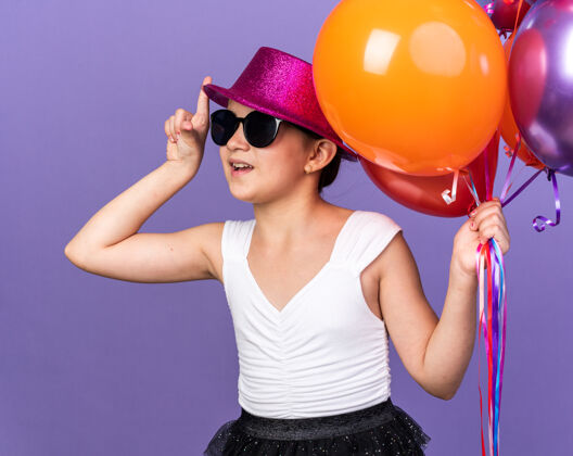 氦惊讶的年轻白种女孩戴着太阳眼镜 戴着紫色的派对帽 手里拿着氦气球 孤立地指着紫色的墙上的复制空间拿着年轻白种人