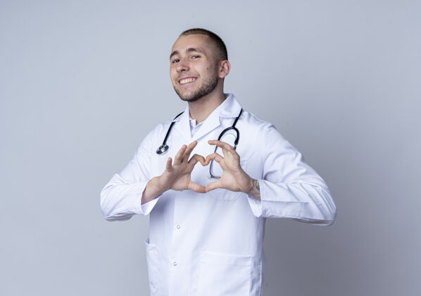 人面带微笑的年轻男医生 身穿医用长袍 脖子上戴着听诊器 在白墙上做着孤立的心脏标志白长袍人