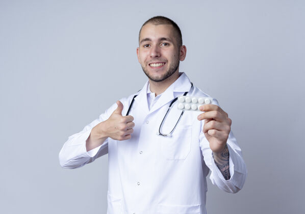 长袍面带微笑的年轻男医生 穿着医用长袍 脖子上戴着听诊器 手里拿着一包药片 在白色的墙上孤立地竖起大拇指医学感情表演