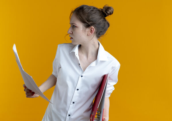 金发年轻严肃的金发俄罗斯女孩拿着文件夹和纸页看着旁边文件背景橙色
