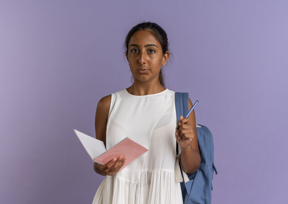 背看着镜头年轻的女学生背着书包拿着笔 笔记本在紫色的背景上笔女学生紫色