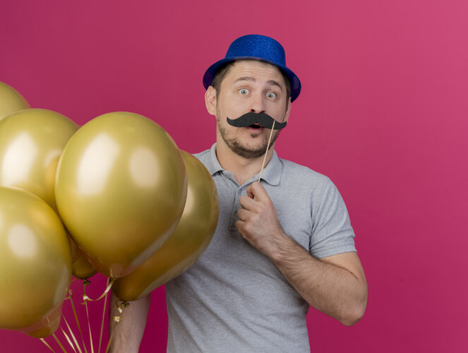 棍子惊讶的年轻人戴着蓝色的帽子拿着气球 假胡子插在粉红色的棍子上气球穿惊喜