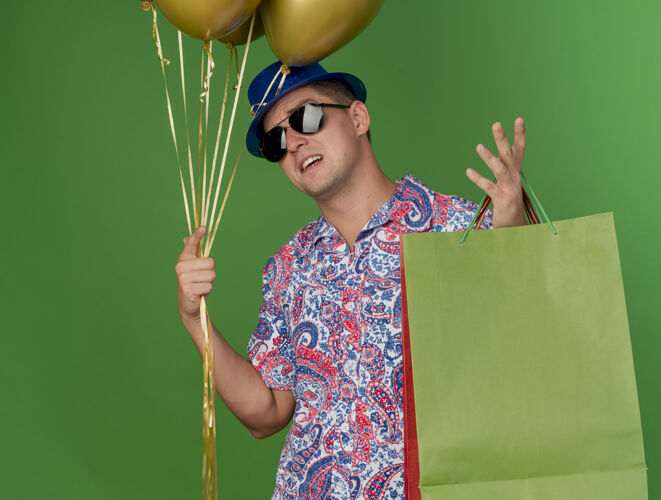 戴着戴着蓝色帽子和眼镜 拿着气球和礼品袋 摊开手在绿色上孤立的年轻人党气球聚会年轻人