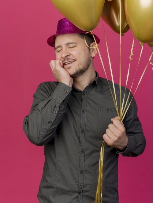 帽子高兴的年轻人闭着眼睛戴着粉红色的帽子拿着气球把手放在脸颊上孤立的粉红色眼睛戴着封闭