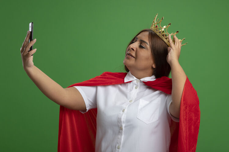 超级英雄高兴的中年女超人戴着皇冠自拍把手放在皇冠上隔离在绿色穿请皇冠