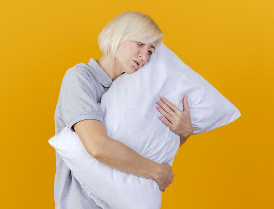 公民脸色苍白的年轻金发病女人抱着枕头 看着橙色墙上孤立的一面人疾病枕头