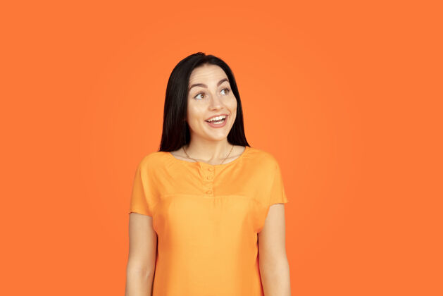 女性橙色工作室背景上的白种人年轻女子肖像穿着衬衫的漂亮深色女性模特人类情感的概念 面部表情 销售 广告广告空间看起来梦幻 微笑广告一半黑发