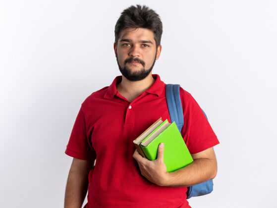 背包年轻的留着胡子的学生 穿着红色马球衫 背着背包 拿着笔记本 站在白色背景下 带着严肃自信的表情看着相机笔记本表情学生