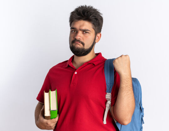 拳头年轻的留着胡子的学生 穿着红色马球衫 背着背包 拿着笔记本 严肃地看着镜头 握紧拳头站在白色背景上衬衫严肃小伙子