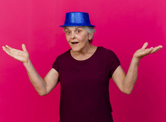 帽子惊讶的戴着派对帽的老妇人在粉红色的墙上张开双手穿人党