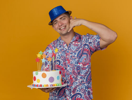蛋糕带着微笑的年轻人戴着蓝色帽子拿着蛋糕 在橘子上做着打电话的手势穿上电话帽子