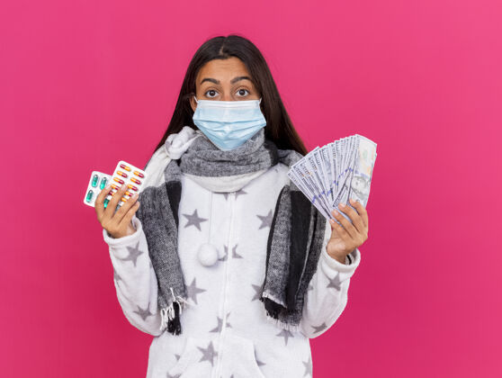 药片惊讶的年轻生病的女孩戴着医用口罩戴着围巾拿着药片拿着现金隔离在粉色上现金医疗粉红色