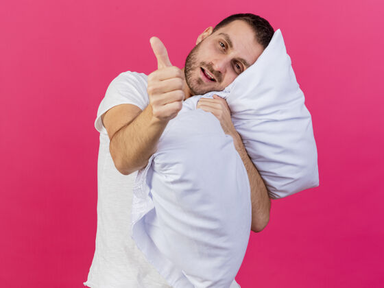 倾斜歪着头的年轻病夫抱着枕头 把拇指孤立地竖在粉红色的床上拥抱疾病粉色