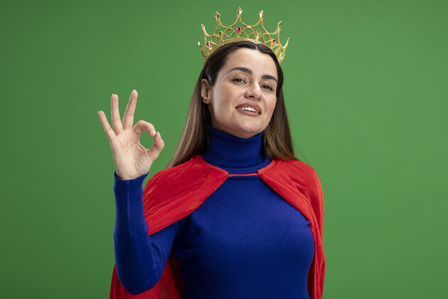 超级英雄戴着王冠的年轻超级英雄女孩很高兴在绿色上显示出良好的姿态拜托好的手势