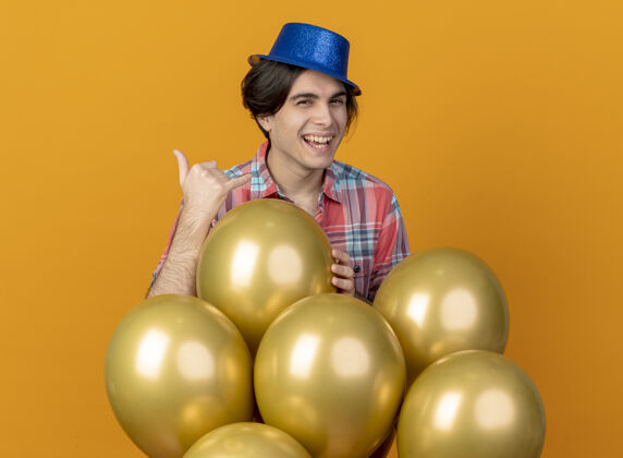表情快乐的帅哥戴着蓝色的派对帽 站在橙色的墙上 拿着氦气球做手势叫我人市民欢乐