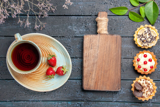 叶子顶视图茶托上的一杯茶和草莓干花枝馅饼叶子和深色木桌上的砧板剁碎酸的木头