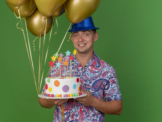 抱着带着微笑的年轻人戴着蓝色帽子拿着气球和蛋糕隔离在绿色戴着绿色小伙子