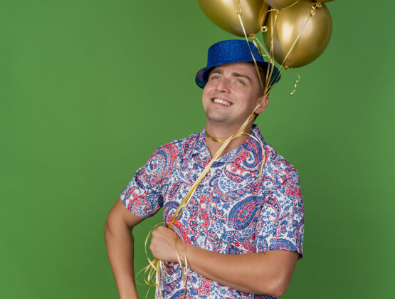 小伙子微笑着的年轻人戴着蓝色帽子抬头看 手里拿着绑在脖子上的气球孤立在绿色的绿色围着穿