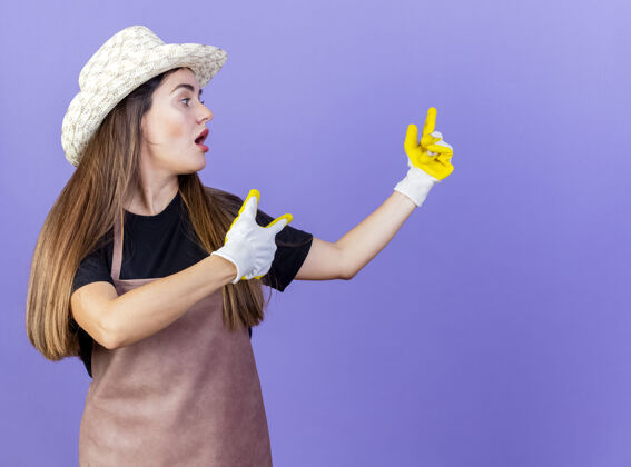 壁板惊讶美丽的园丁女孩穿着制服戴着园艺帽和手套点在一边孤立的蓝色园艺点手套
