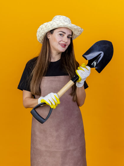 穿微笑美丽的园丁女孩穿着制服 戴着园艺帽 戴着手套 手持铁锹 孤立在橘子上橙色手套制服