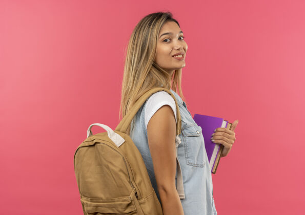 漂亮高兴的年轻漂亮的学生女孩穿着背袋站在侧面图举行孤立的粉红色墙上的书人粉色脸