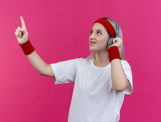 壁板微笑的年轻运动女性戴着耳机戴着护腕戴着头带 看着粉色墙壁上孤立的一面头带微笑人