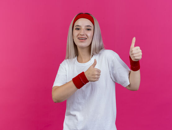女人快乐的年轻运动女性戴着背带戴着头带和腕带两只手竖起大拇指孤立在粉红色的墙上欢乐头带佩戴