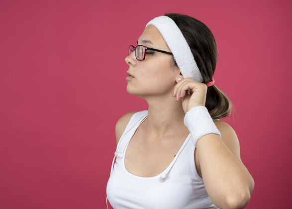 耳朵自信的年轻运动女性戴着眼镜 头戴耳机 戴着头带和腕带 抱着耳朵 看着粉色墙上孤立的一面眼镜人年轻人