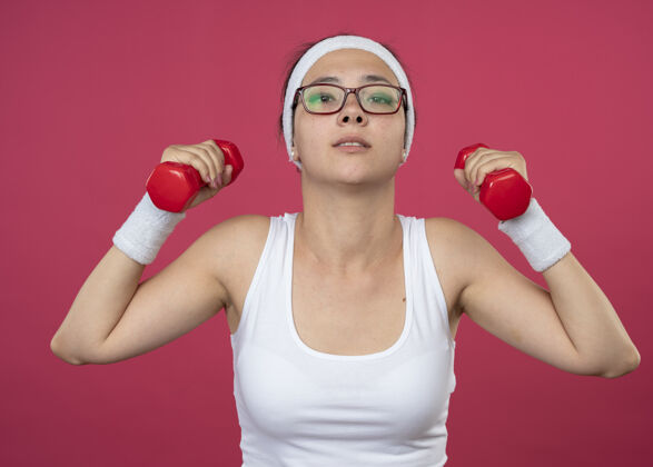 自信戴着眼镜 戴着头带和腕带的自信的年轻运动女性 在粉红色的墙上孤立地举着哑铃戴眼镜人