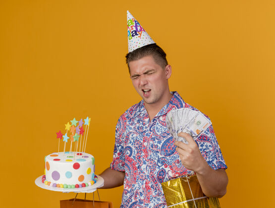 家伙快乐的年轻人 戴着生日帽 闭着眼睛 手里拿着生日礼物 蛋糕和现金被隔离在橘子上生日帽子年轻
