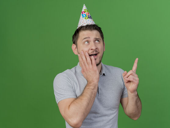 男人惊讶的年轻人戴着生日帽把手放在脸颊点在绿色上孤立起来生日帽子年轻