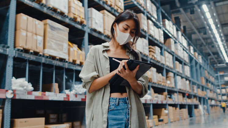 健康年轻的亚洲女商人经理戴着口罩仓库使用数码平板电脑检查库存航运工人货架