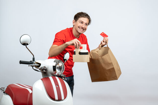 微笑身着红色制服 面带微笑的送货员站在滑板车旁 手持白色背景的银行卡订单自行车滑板车男