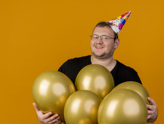 成人戴着生日帽 戴着眼镜 面带微笑的成年男子站在橙色墙壁上 站着氦气球脸眼镜人