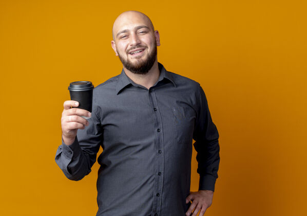 感觉微笑着的年轻秃顶呼叫中心的男人 拿着塑料咖啡杯 把手放在腰上 隔离在橙色的墙上杯子电话人