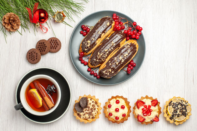 圣诞在灰色的盘子上俯瞰巧克力eclairs和醋栗柠檬肉桂茶馅饼饼干和松树叶子与圣诞玩具在白色的木制地面食物松树盘子