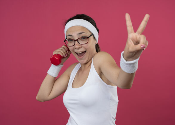 胜利戴着眼镜戴着头带和腕带的快乐的年轻运动女性手持哑铃 手势胜利手势孤立在粉红色的墙上哑铃女孩腕带