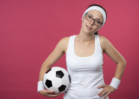 请戴着眼镜 戴着头带和腕带的快乐的年轻运动女性把手放在腰上 把球孤立地举在粉红色的墙上运动拿着头带