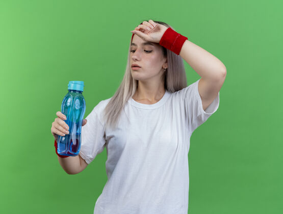 戴自信的年轻运动女性戴着背带 戴着头带和腕带 把手放在额头上 看着隔离在绿色墙上的水瓶年轻人女孩女人